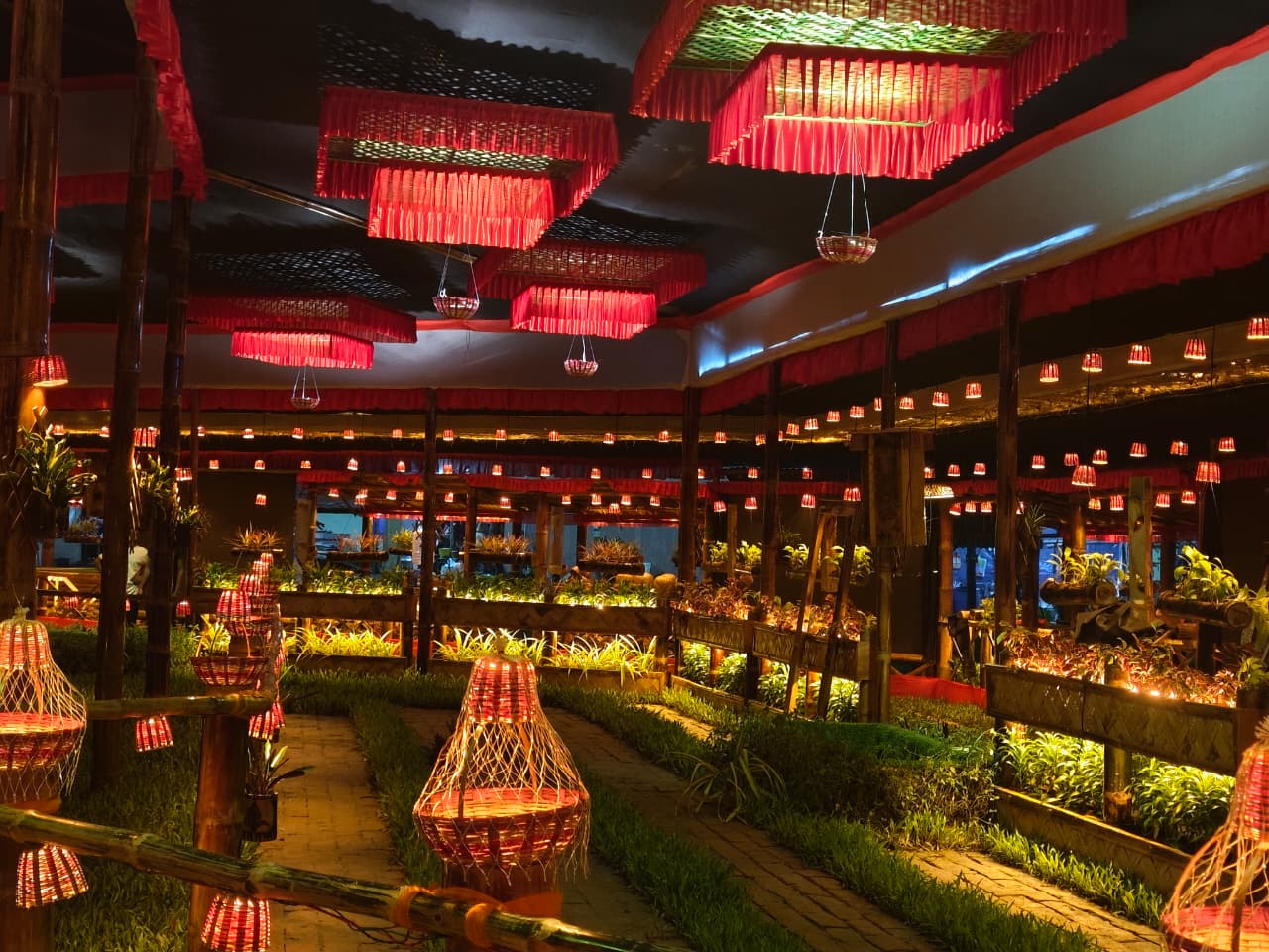 Inside the Museum of the Future — red woven chandeliers overhead, woven basket lanterns glowing along bamboo fences, garden beds with living plants between brick pathways